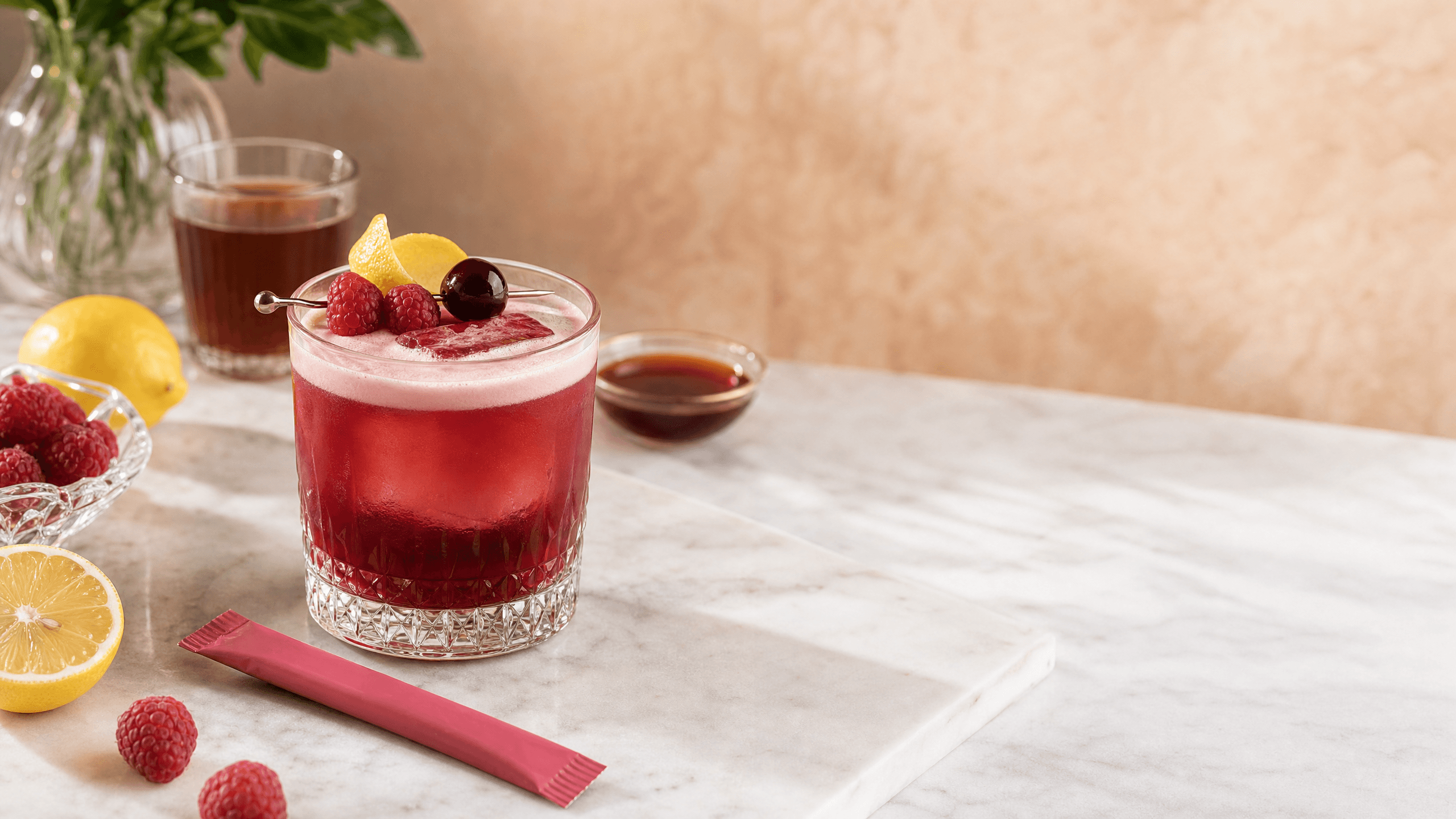 A raspberry kava sour in a rocks glass with foam, lemon peel, raspberries, and a dark cherry garnish